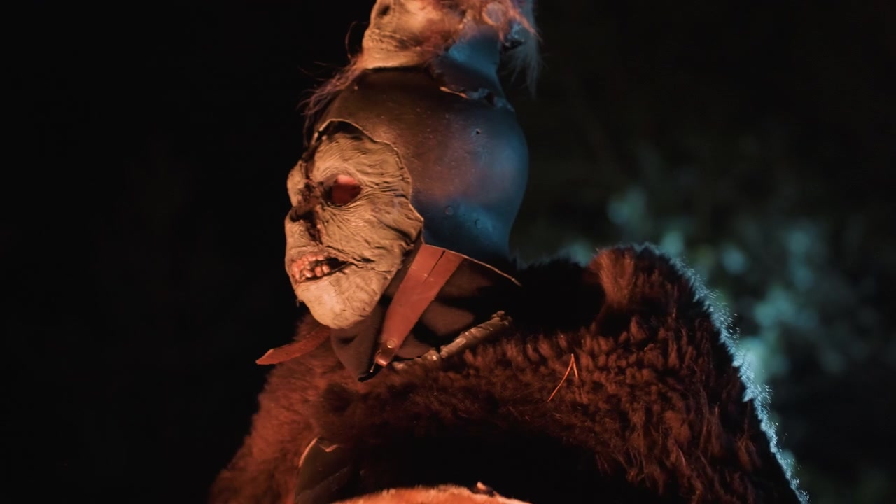 Low-angle close-up shows two stacked carved skull masks mounted on round painted shield or drum, with teal rim light, dark shadows, textured surfaces, fur base, and ominous ceremonial presence