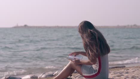 slim long haired lady reads book sitting on sandy beach