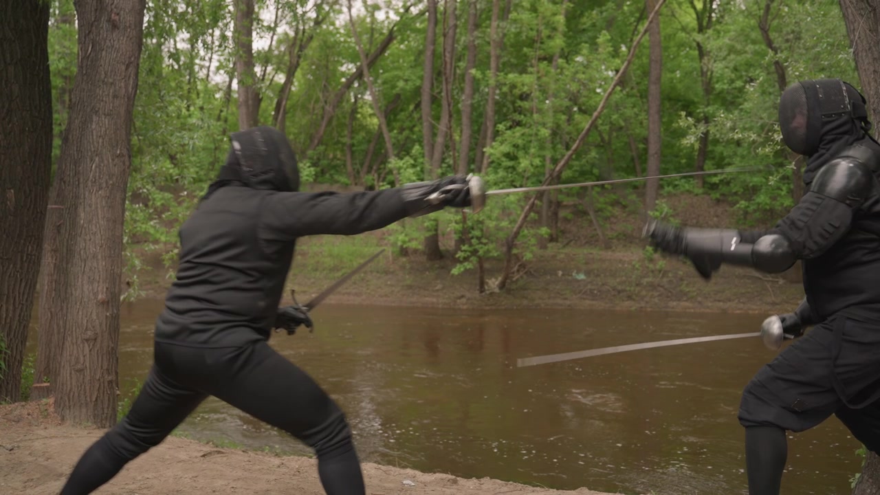 Two people in black fencing gear engage with swords near riverbank surrounded by trees. Their movements are fluid as they parry and thrust demonstrating skill against each other. natural