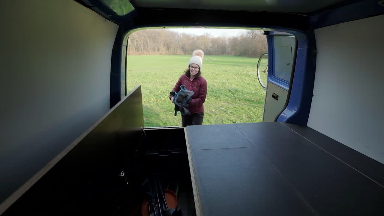A woman walking towards the back of a camper with a chair. Camera looking out the back of the camper van and the women pack up a chair and places it in storage. A couple travelling in a campervan