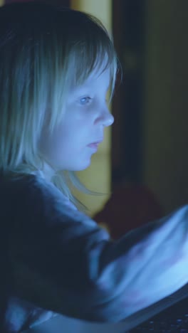 young child focused on using laptop in dimly lit room, illuminated by screen light, showcasing concentration and engagement with technology