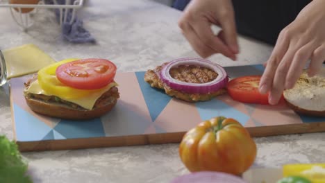 hands layering tomato and bell pepper slices onto a burger patty and cheese on a colorful cutting board during a homemade sandwich prep session.