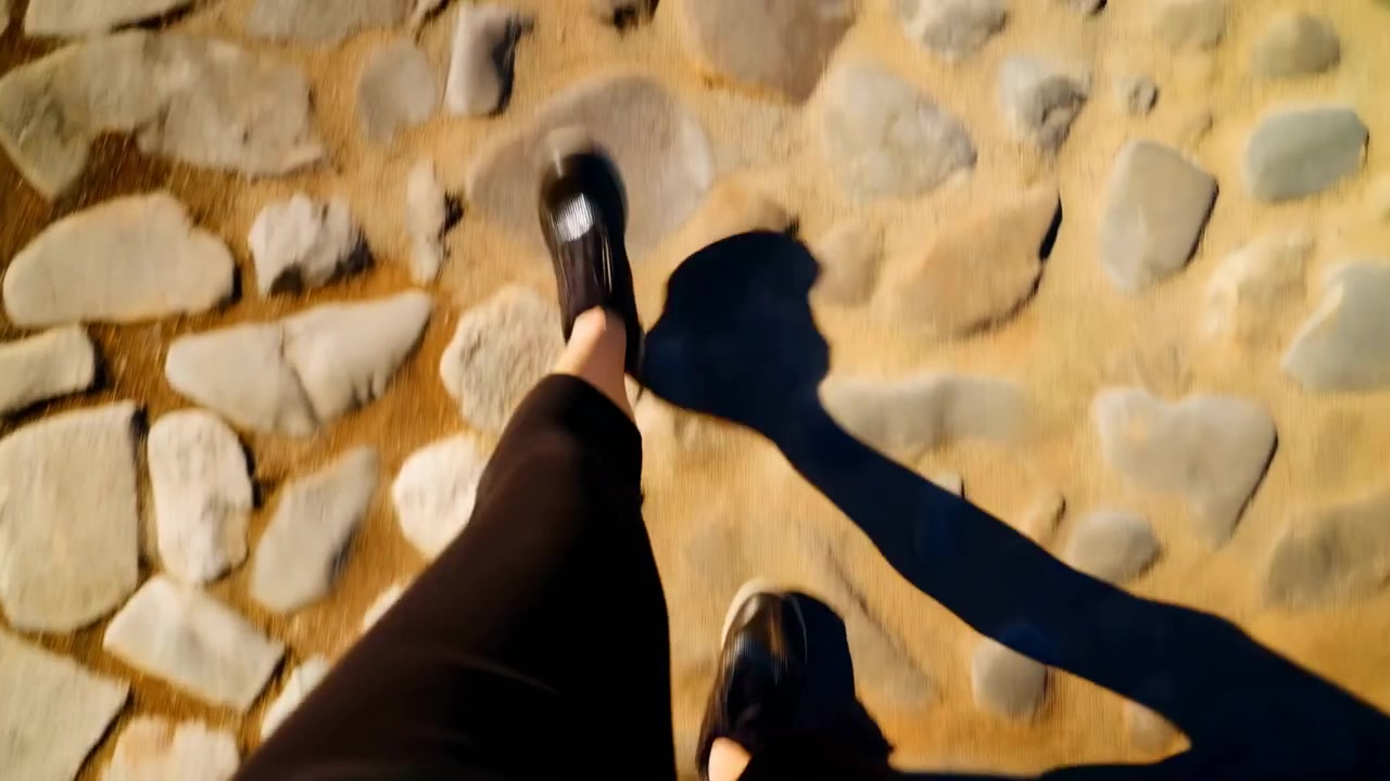 First-person walking scene on sunlit cobblestone walkway, showing glossy sneaker, cropped black pants, long shadow, textured stone surface, and warm outdoor daylight with urban travel mood