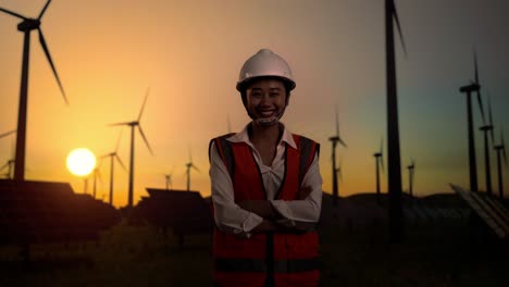 asian female engineer in a helmet smiling and posing with arms crossed while standing in front of wind turbines rotating at sunset