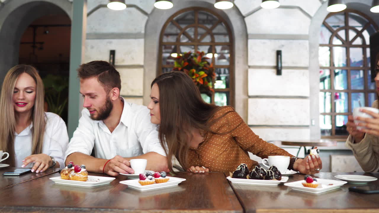 dos chicas sentadas en un café mirando el teléfono y discutiendo lo que vieron. un tipo se acerca y muestra una foto graciosa en su teléfono