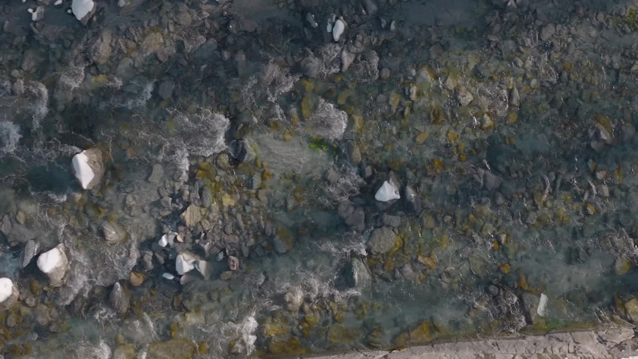 toma aérea lenta de arriba hacia abajo de un pequeño y lindo río poco profundo en un pueblo suizo que fluye junto a las rocas y debajo del puente en un hermoso día soleado de invierno con nieve a un lado de la orilla del río