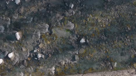 slow aerial top down shot of cute small shallow river in swiss village flowing alongside rocks and under bridge on beautiful sunny winter day with snow on side of river bank