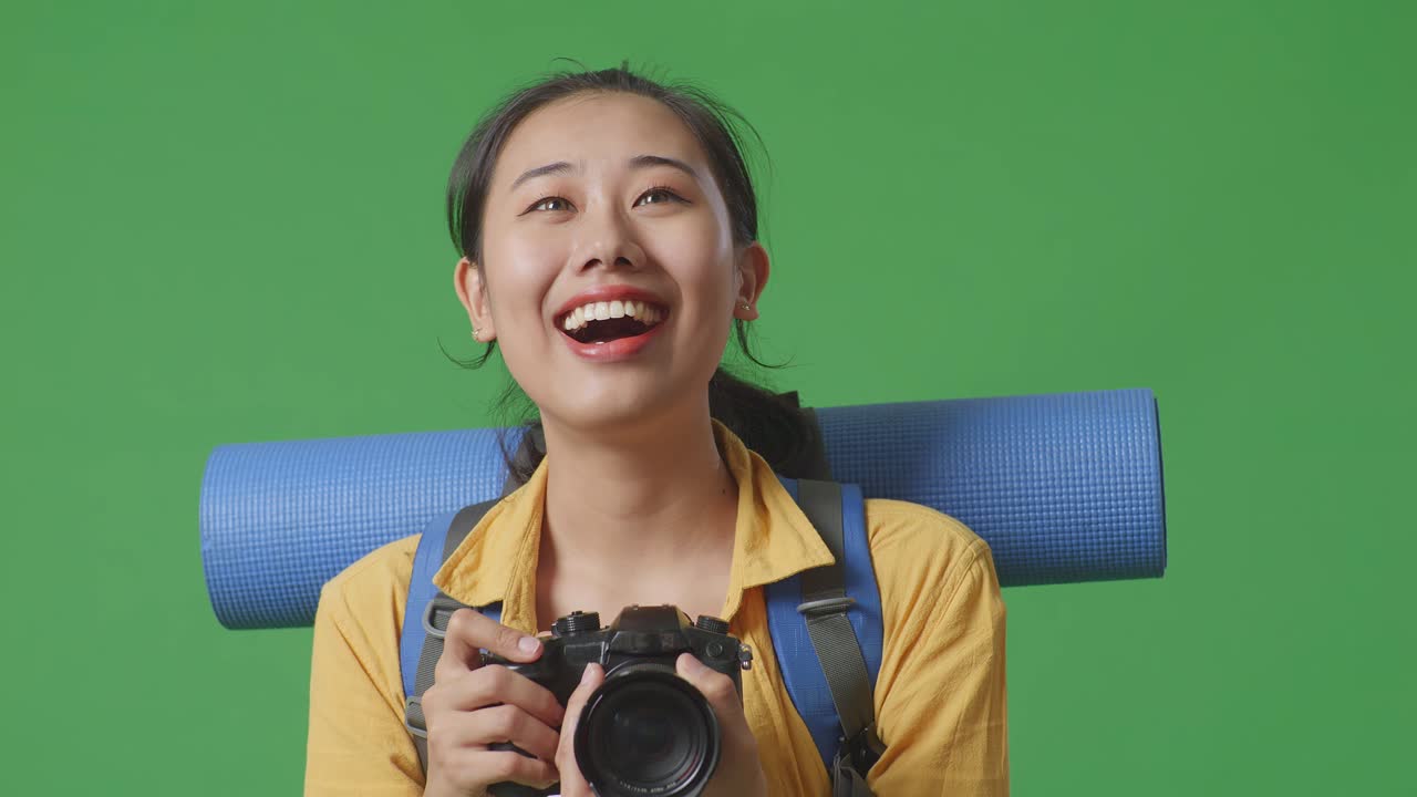 아시아 여성 하이커가 산악 배을 들고 웃으며 손에 카메라를 들고 스튜디오에서 녹색 화면 배경에 서서 주위를 둘러보고 있습니다.