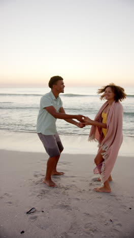 vertical video: couple dancing on beach, enjoying sunset wrapped in blanket, having fun