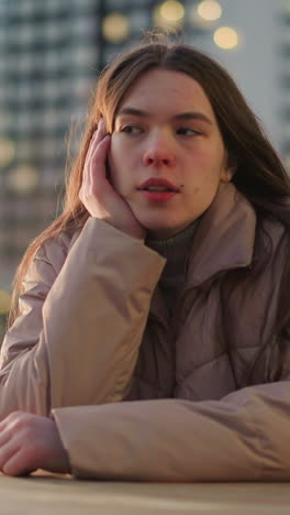 un primer plano de una mujer joven con una chaqueta de melocotón, perdida en pensamientos profundos mientras descansa la mejilla en su mano. sus ojos están cerrados, y el fondo urbano borroso