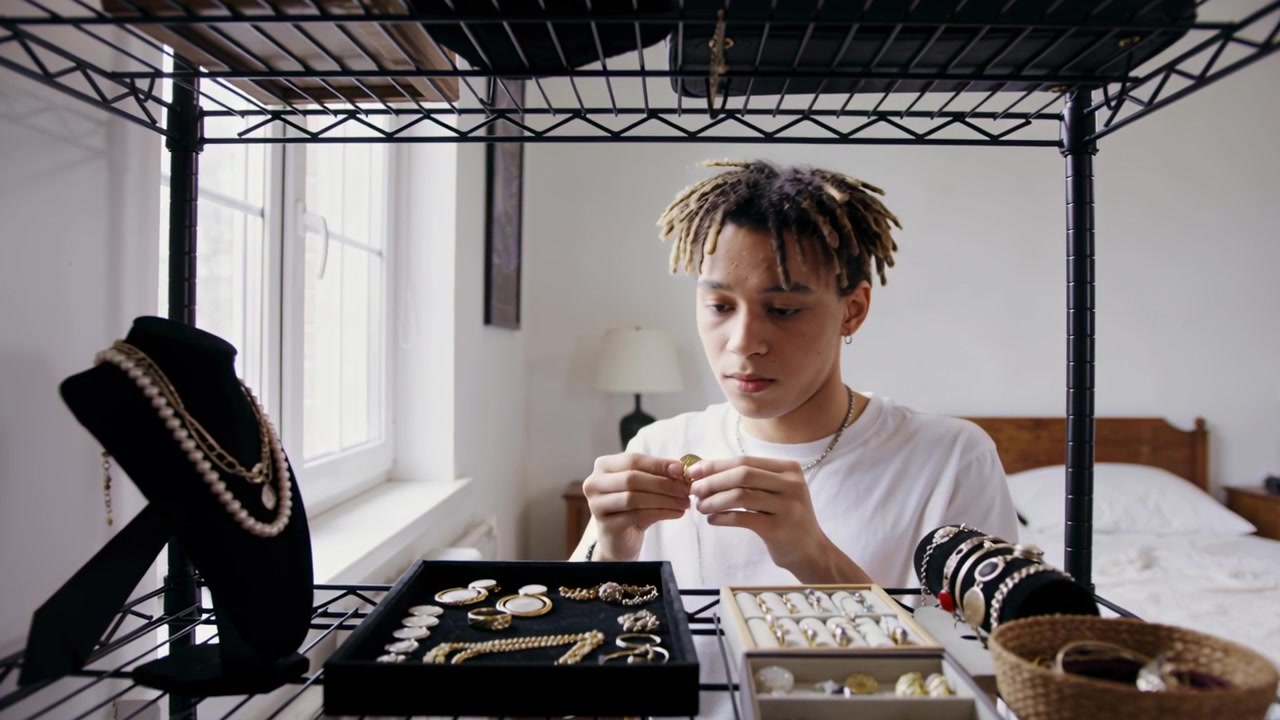 Lifting necklace Black teen in white tee inspecting clasp and organizing jewelry on wire shelving
