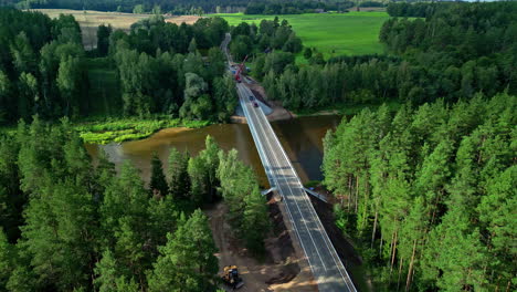 Aerial-drone-shot-over-river-flowing-through-a-forest-alongside-a-bridge-on-a-cloudy-day