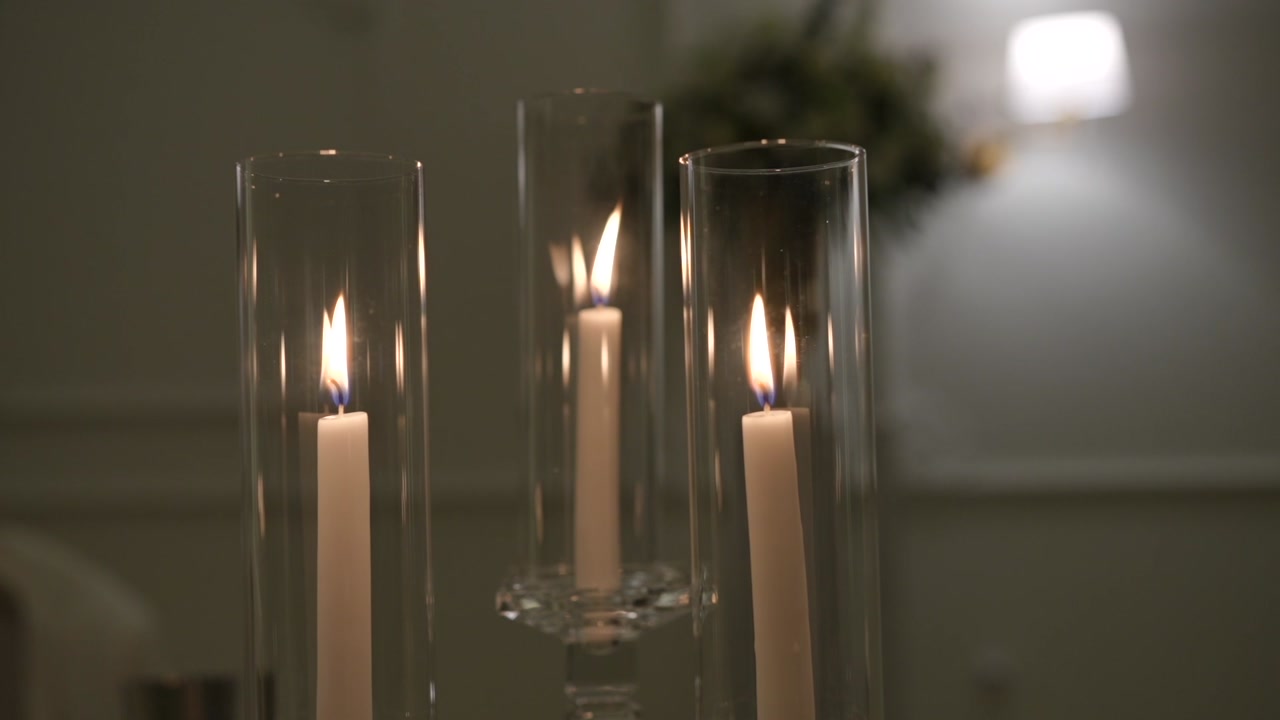 Three lit taper candles inside tall clear glass cylinder hurricane holders on a table, soft bokeh sconce and greenery in background