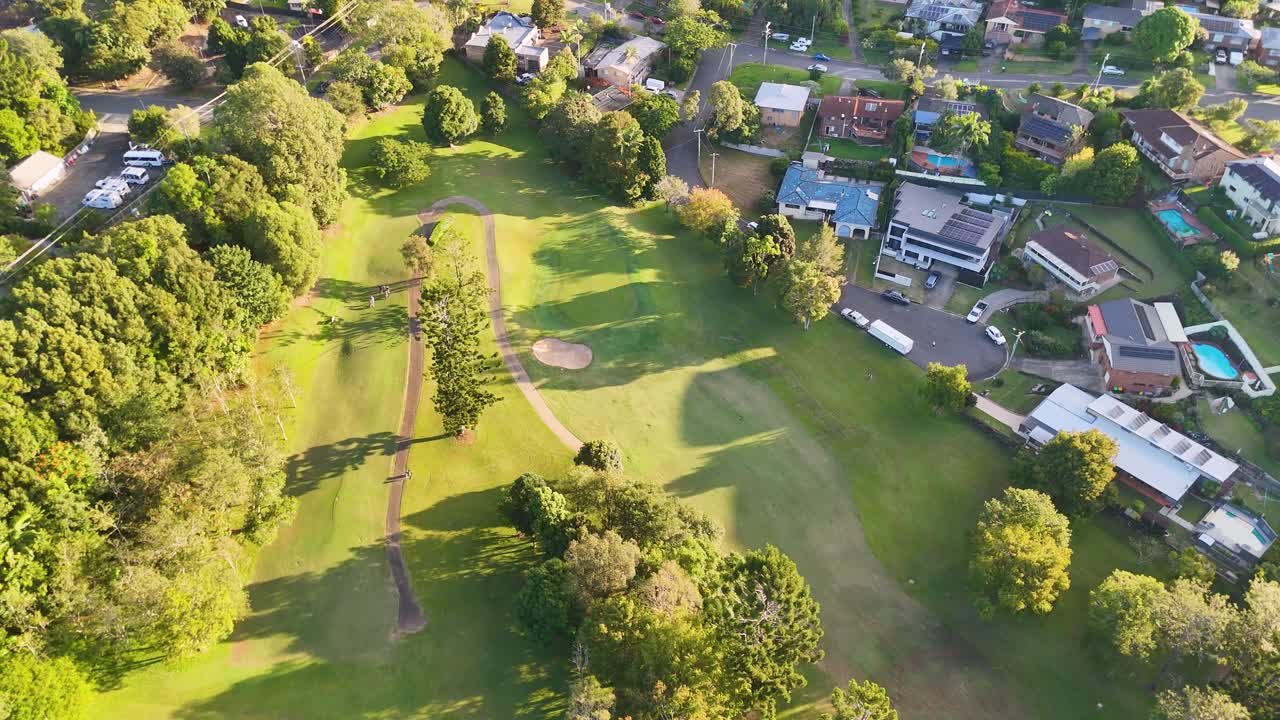 Drone footage captures a lush golf course adjacent to a residential neighborhood under bright sunlight