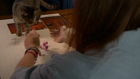 woman rolls pink rpg dice on game table near pet cat