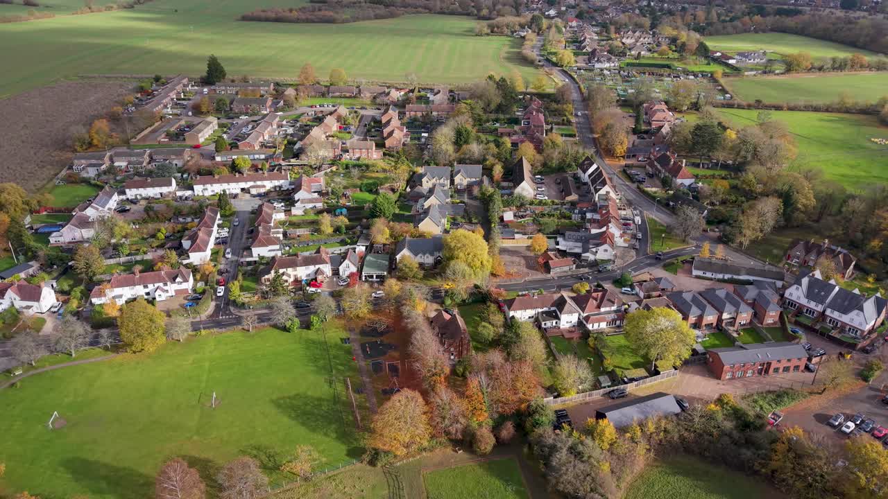 Drone reverses and rises over High Wych village, Essex, revealing its small size, surrounded by fields and trees. Captured under broken clouds, sunlight highlights parts of the rural autumn landscape
