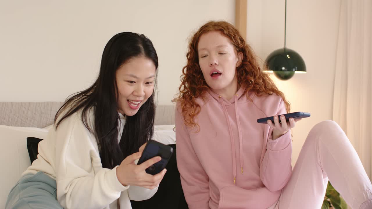 Diverse amiche con felpa e felpa rosa, sedute su cuscini di un divano basso, una indica il telefono e confrontano.