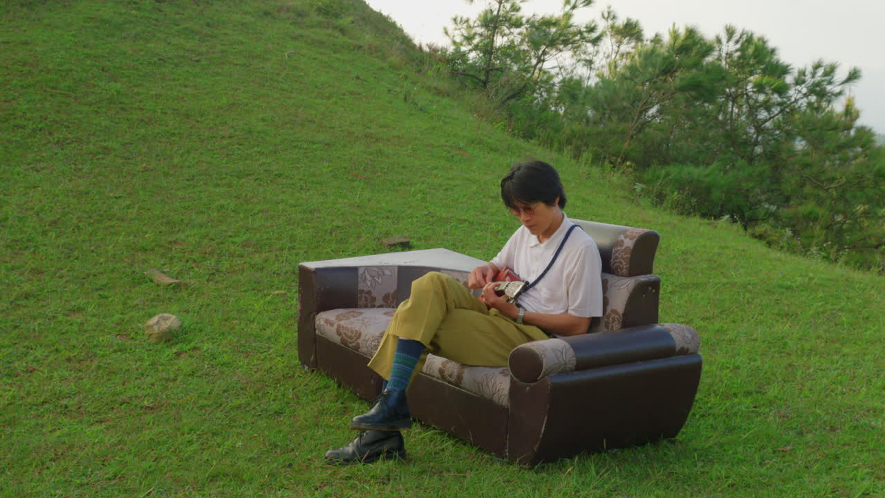 젊은 중국 예술가 한 명 은 야외 에서 가죽 소파 소파 에 앉아 유클레 기타 를 연주 하고 있다.