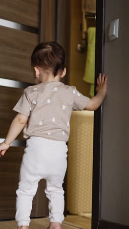 cute toddler stands near the door. kid pushes the door and looks how it opens ajar. low angle view. vertical video