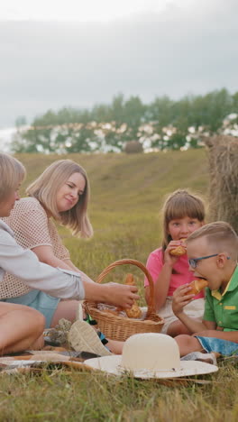 familia sentada en la hierba disfrutando de pasteles frescos de la canasta de picnic, niño en camisa verde come inmediatamente mientras que la niña en camisa rosa elige cuidadosamente su elección