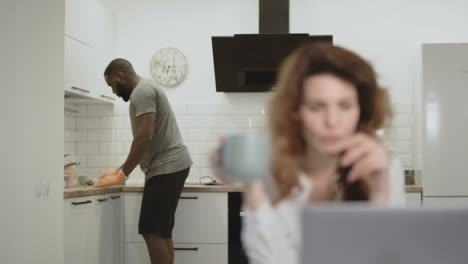 Hombre-Negro-Limpiando-La-Mesa-En-La-Cocina-Abierta.-Mujer-Bonita-Bebiendo-Café-Por-La-Mañana