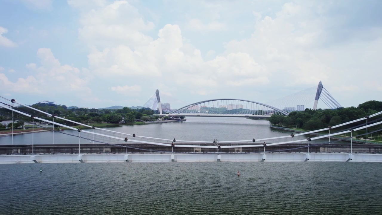 Boční záběr dronem zachycující vyřazený most monorailu Putrajaya s mostem Seri Saujana viditelným v pozadí nad klidnou nábřežní scenérií.