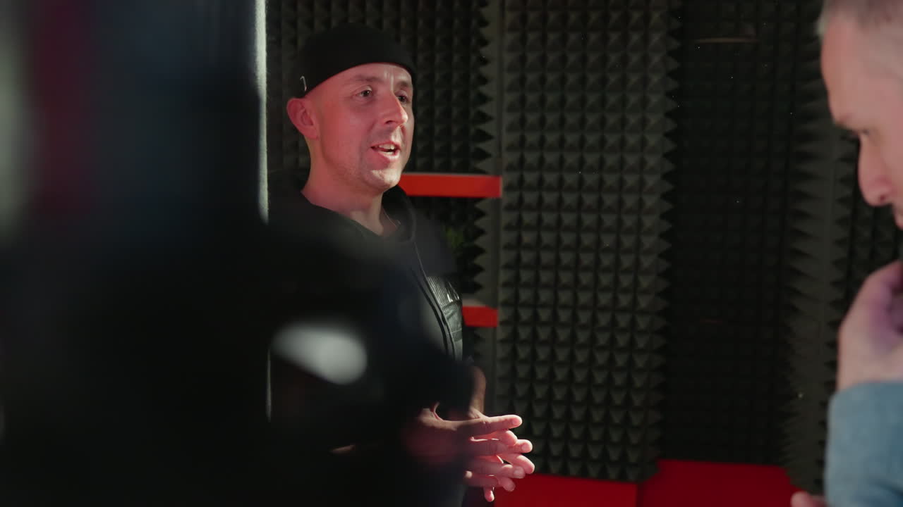 A man in a black shirt and cap enthusiastically talks to another person, barely visible, set against a black soundproof room with striking red details, highlighting a moment of vibrant communication