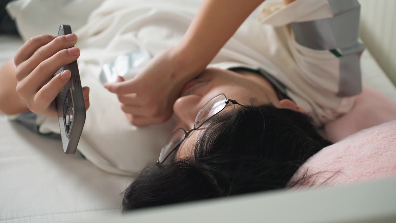 Asian woman student lying on bed, groggy morning scrolls smartphone while adjusting glasses, soft pillow and blanket, warm window light, candid domestic portrait conveying tired routine and quiet
