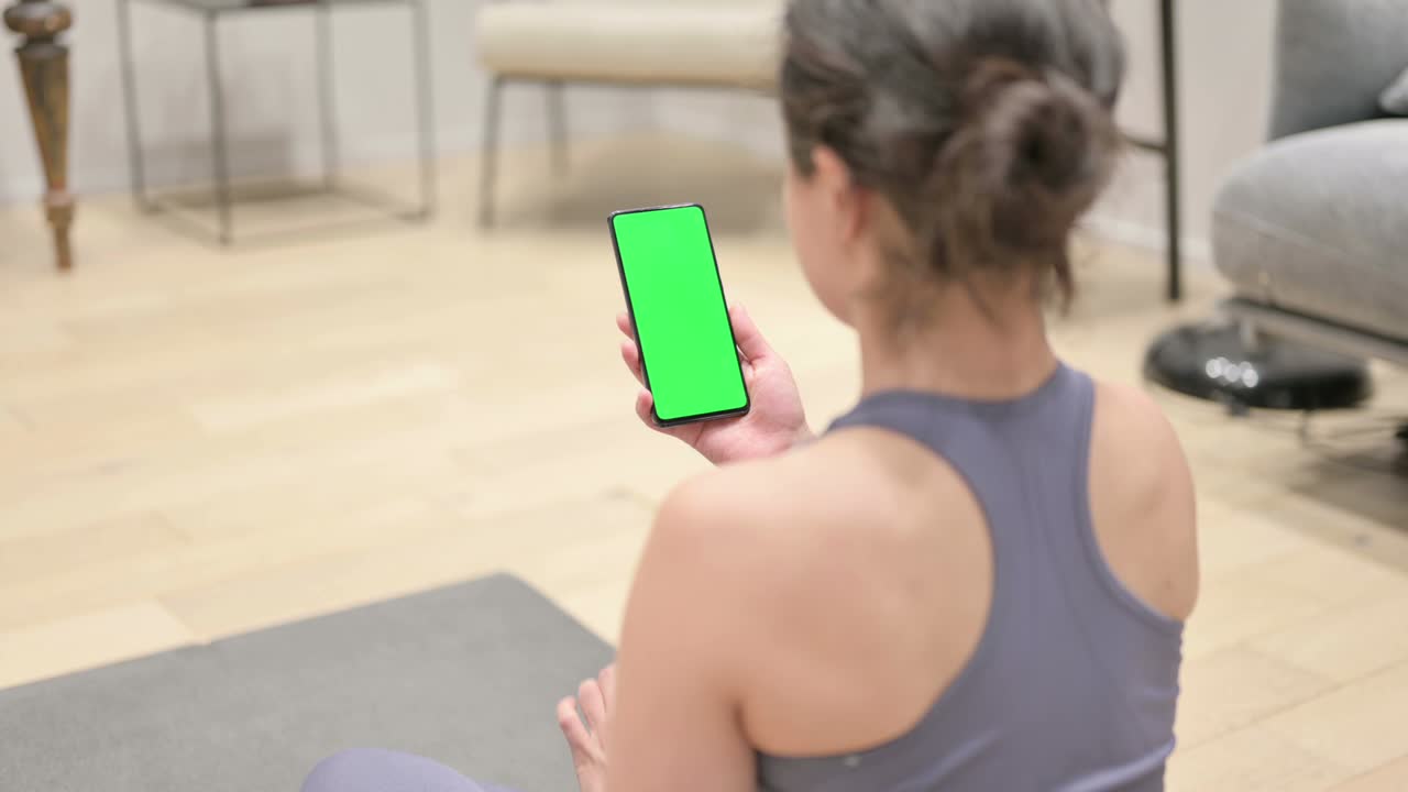 vista trasera de una mujer india usando un teléfono inteligente con pantalla cromada en una alfombra de yoga