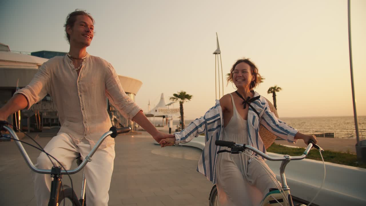 Happy guy and girl riding bicycles on the beach looking at each other and holding hands near the sea at sunrise in summer. Happy couple ride bicycles on the beach with palm trees near the sea in summer