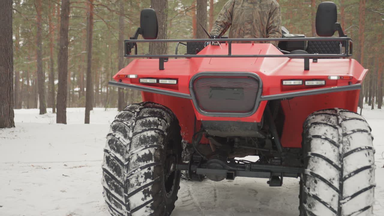 Un ATV rosso in una pineta innevata durante una corsa fuoristrada estrema, con il pilota che manovra nella neve compattata. Primo piano dal basso delle ruote anteriori e della robusta griglia, pneumatici con battistrada profondo che sollevano la neve, sospensioni robuste.