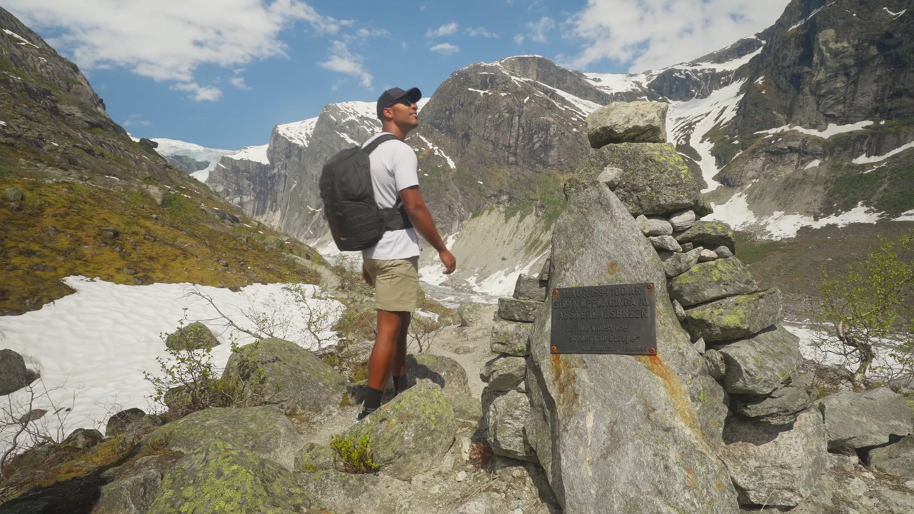 Due giovani escursionisti in forma camminano lungo un sentiero di montagna circondati da cime innevate, terreno roccioso e segnaletica per il ghiacciaio Austerdalsbreen in Norvegia.