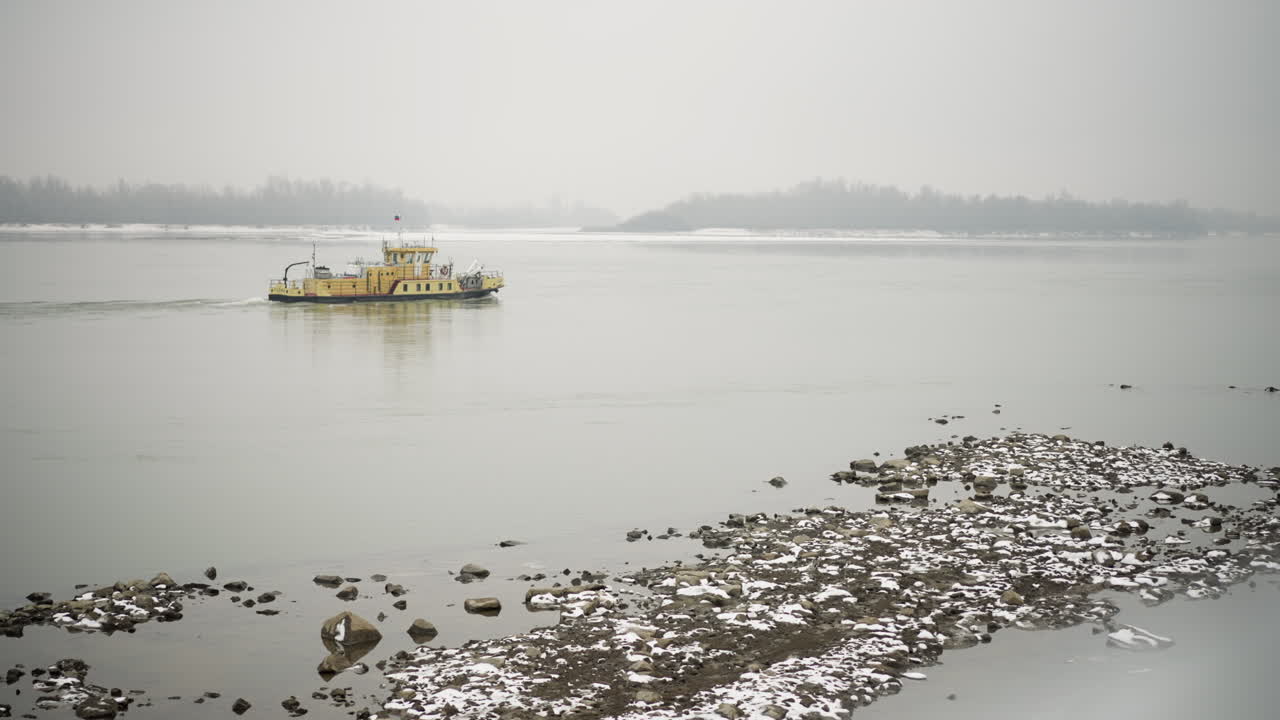 노란색 배는 바위 해안 근처의 안개 강에서 조용히 항해하며, 나무의 반사와 멀리 있는 눈 인 땅, 평온한 겨울 물과 가운 안개에 둘러싸여 평화로운 분위기를 만니다.