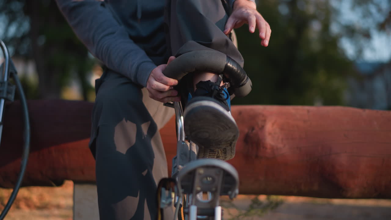 회색 후디와 어두운 바지를 입은 젊은이가 밖에서 나무 에 앉아 활 기 장비를 착용하는 것에 집중하고 있습니다. 배경에는 흐릿한 초록색이 있습니다.
