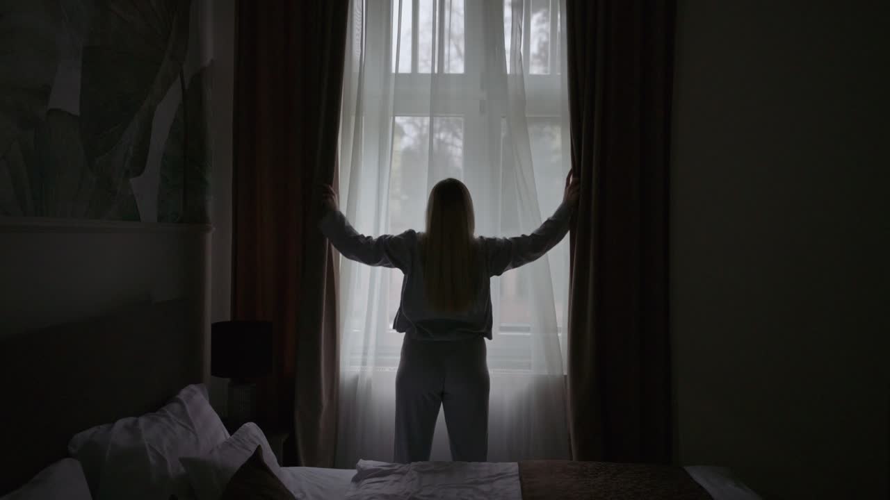 Silhouette di una donna in piedi in una camera da letto mentre apre le tende per far entrare la luce del mattino.