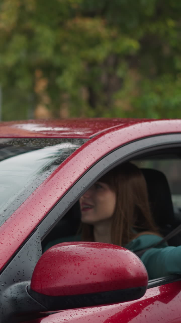 Lady drives red car with wipers cleaning windshield reflecting trees on rainy day. Happy woman in green jacket puts hand out of window to catch rain slow motion