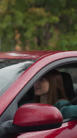 una mujer conduce un coche rojo con limpiaparabrisas que limpia el parabrisas que refleja los árboles en un día de lluvia. una mujer feliz con una chaqueta verde pone la mano fuera de la ventana para atrapar la lluvia en cámara lenta