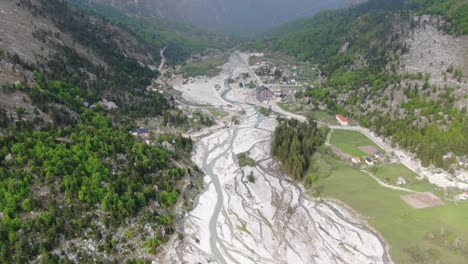 Drone-view-in-Albania-flying-in-the-alps-showing-green-forest-on-a-valley-surrounded-by-mountain-with-snowy-peaks-and-rivers-flowing-in-Valbon?