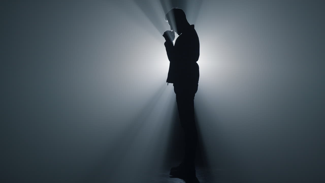 silueta de un creyente orando en un espacio oscuro. un hombre serio de pie con las manos cruzadas.
