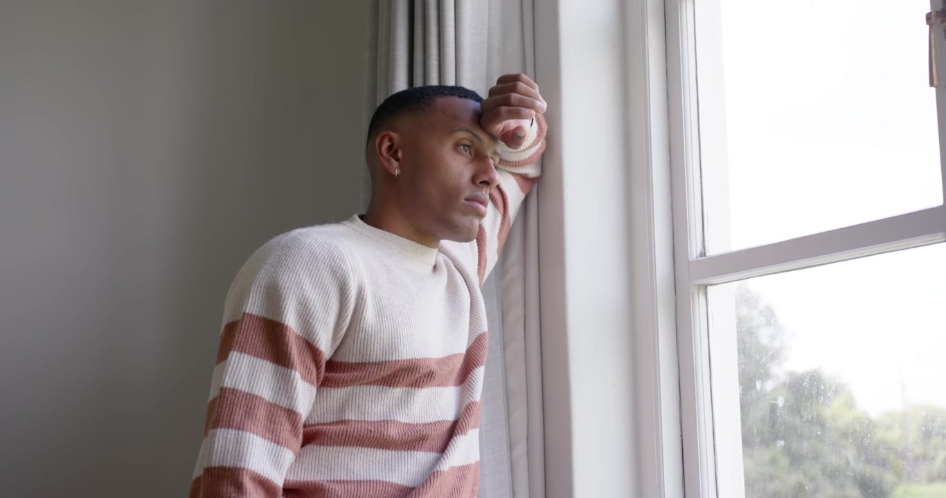 African American man entering frame, leaning on window at home, gazing outside and reflecting