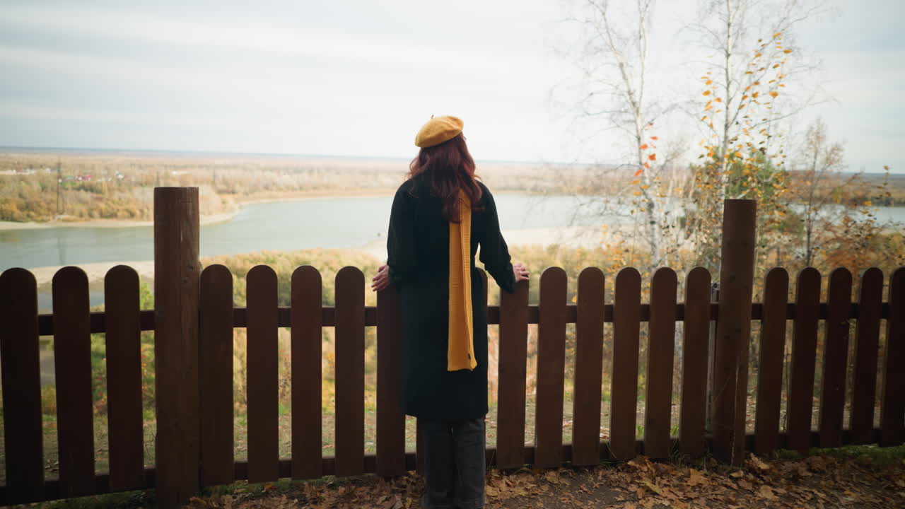 검은 코트를 입은 젊은 여성이 나무 울타리를 향해 고, 건조한 나무 잎자루와 멀리 있는 강과 함께 숨을 쉬는 자연 장면을 즐기면서 손을 고 있습니다.