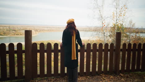 back view of a young woman in a black coat walking towards a wooden fence, resting her hands on it while enjoying a breathtaking nature scene with dry tree foliage and a distant river