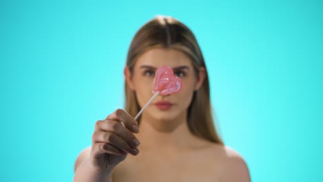 medium shot of a young beautiful woman holding a heart shaped delicious lollipop into the camera in front of turquoise background in slow motion