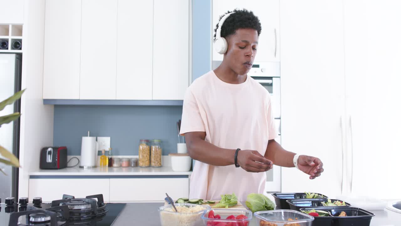 Afroamerikanischer Mann in pinkfarbenem T-Shirt mit weißem Headset stellt Mahlzeiten an einer Kücheninsel mit schwarzen Tabletts zusammen.