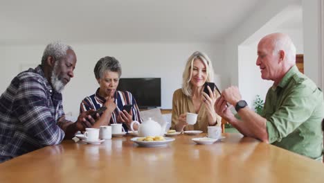 dos parejas mayores diversas sentadas junto a una mesa bebiendo té usando teléfonos inteligentes en casa