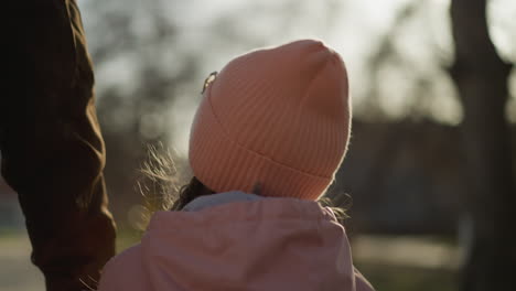 a little girl in a pink cap and jacket, joyfully holding a man's hand while walking outdoors. the girl looks up with a smile, and the warm sunlight creates a happy and serene atmosphere