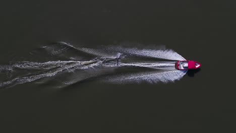 a professional water skier jumping and weaving through the water while being pulled by a red speedboat - aerial shot