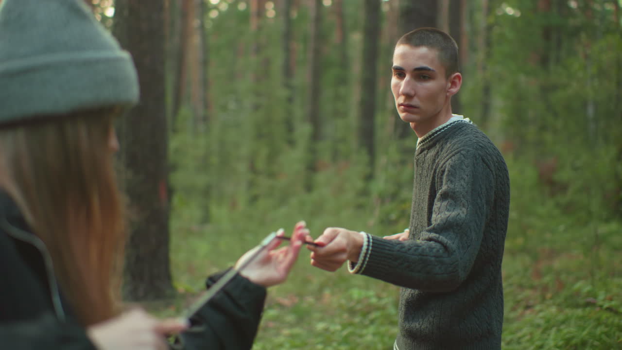 Boy and colleague joining ends of flexible tent pole while setting up camping gear in forest, tent visible in background, surrounded by trees and greenery