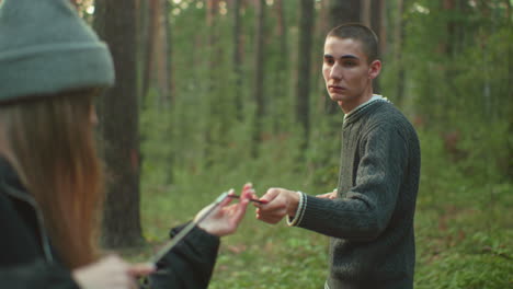 boy and colleague joining ends of flexible tent pole while setting up camping gear in forest, tent visible in background, surrounded by trees and greenery