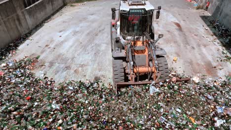 Forklift-excavator-is-grabbing-glass-and-loading-it-to-back-of-an-transporter-truck,-heavy-duty-machine-has-a-long-arm-fitted-with-a-glass-grabbing-attachment,-drone-view,-glass-recycling-concept